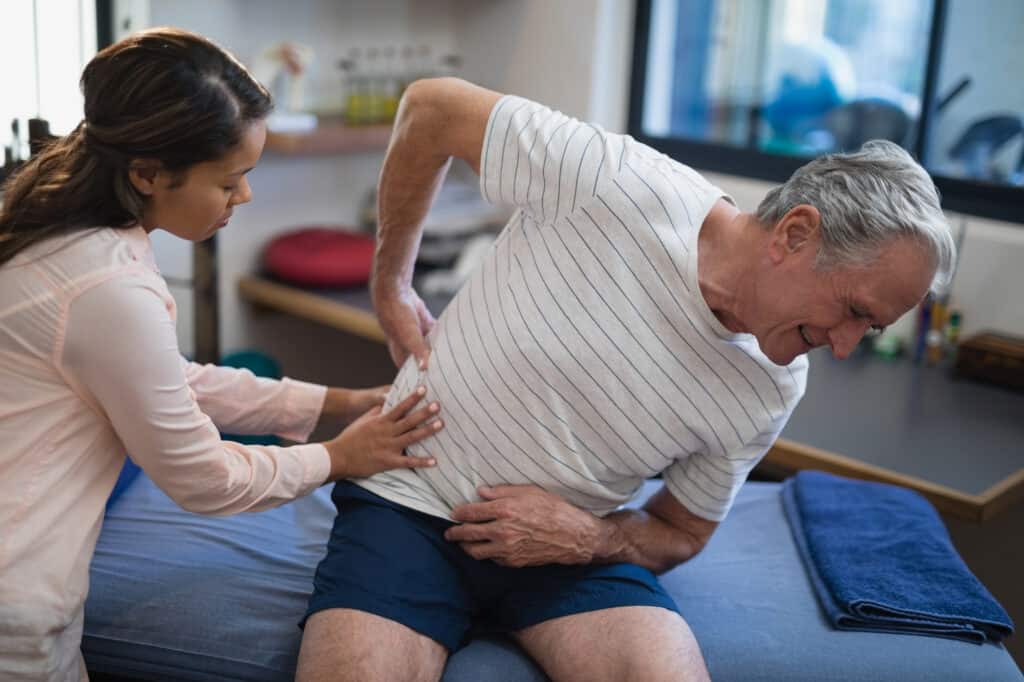 Female therapist examining back of senior male patient sitting on bed at hospital ward